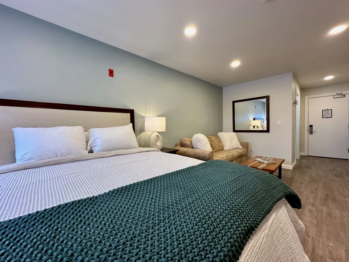 The image features a welcoming studio apartment interior with a plush bed dressed in a green blanket, a comfortable sofa perfect for unwinding, a stylish mirror above the sofa, and a warm bedside lamp. The scene exudes a sense of cozy relaxation, ideal for guests looking to relax after a fun-filled day in the mountains.