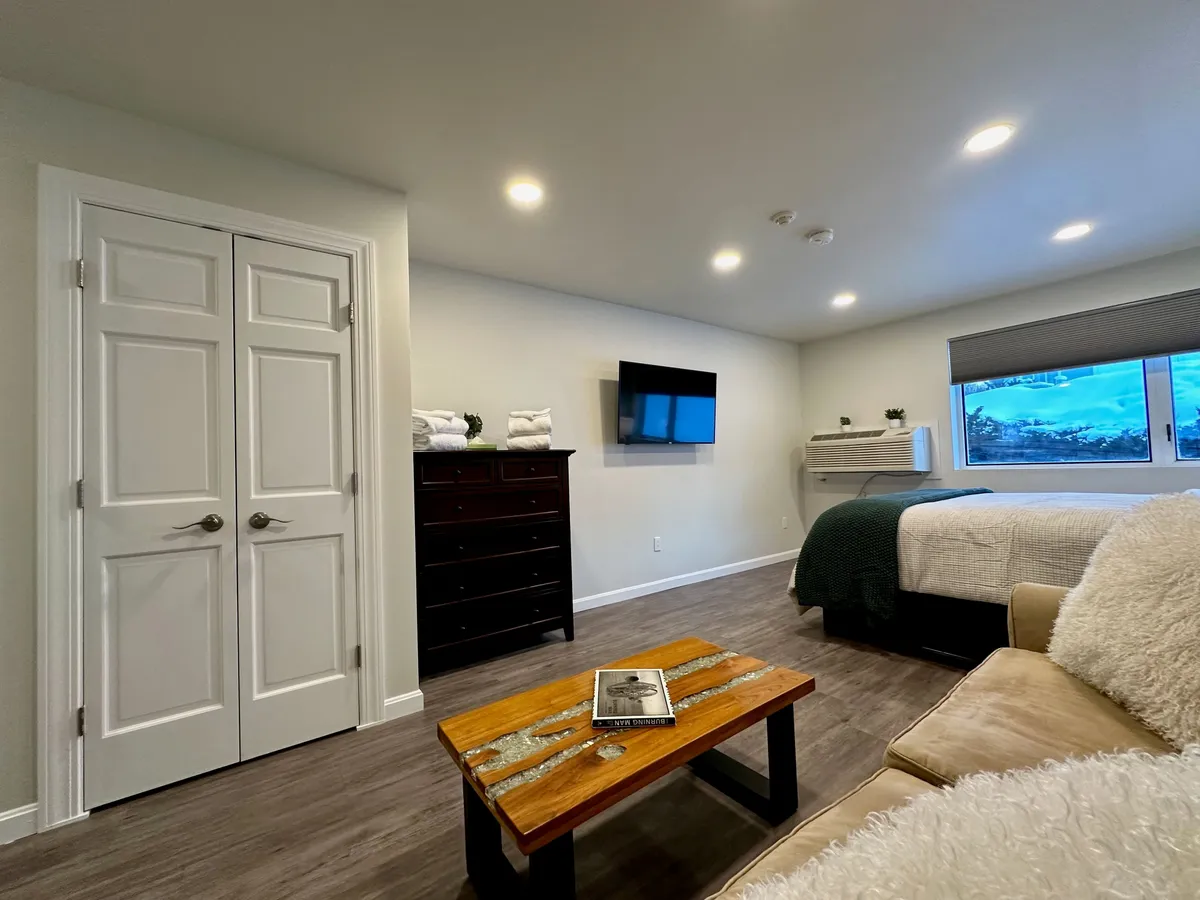 A welcoming interior view of a suite featuring a cozy bed, a television, and a coffee table beside a bright window. The space offers an inviting atmosphere for relaxing with a morning coffee or unwinding after a night out.