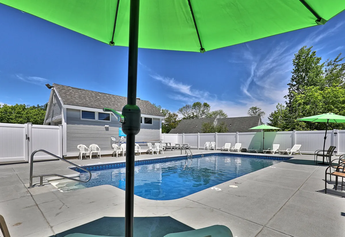 An outdoor swimming pool area surrounded by lush trees, green umbrellas, and lounge chairs. The fenced pool is an inviting spot for relaxation and socializing, with hints of a lively, resort-like atmosphere.