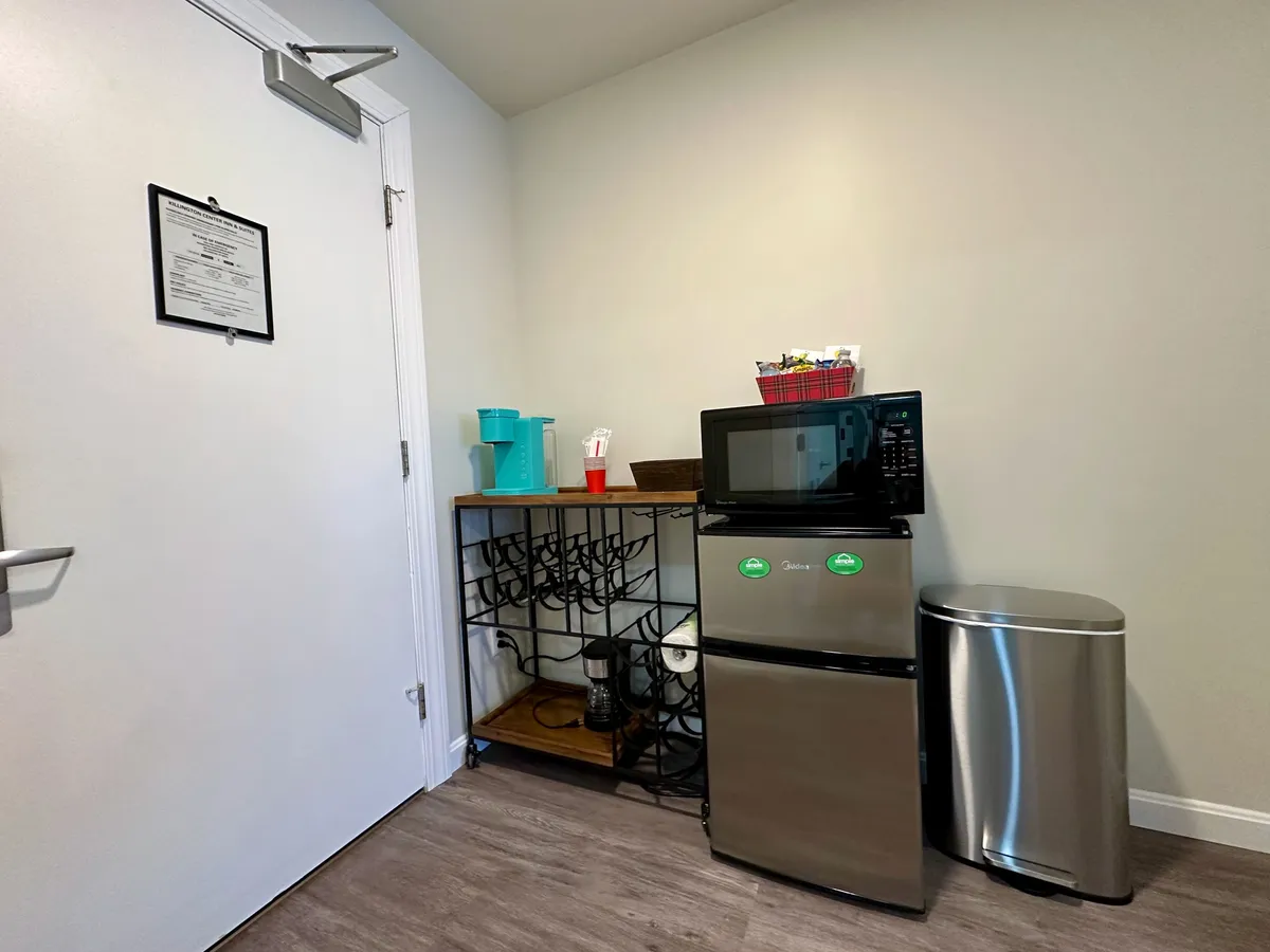 The image shows a well-organized refreshment nook featuring a microwave, a mini-refrigerator, open shelving, and a trash can, all arranged near a doorway. The setting is casual and functional, with everything within easy reach. The lighting and clean layout make the nook inviting for guests to grab a coffee, store drinks, or prepare a quick snack.