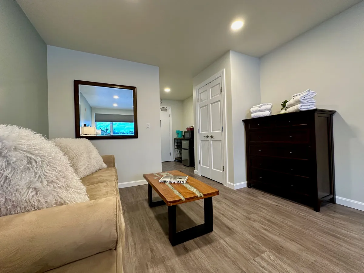 A warmly lit suite living area featuring a plush sofa with soft cushions, a wooden coffee table, a large mirror on the wall, and a dresser topped with neatly folded towels. The inviting arrangement creates a sense of comfort and hospitality.