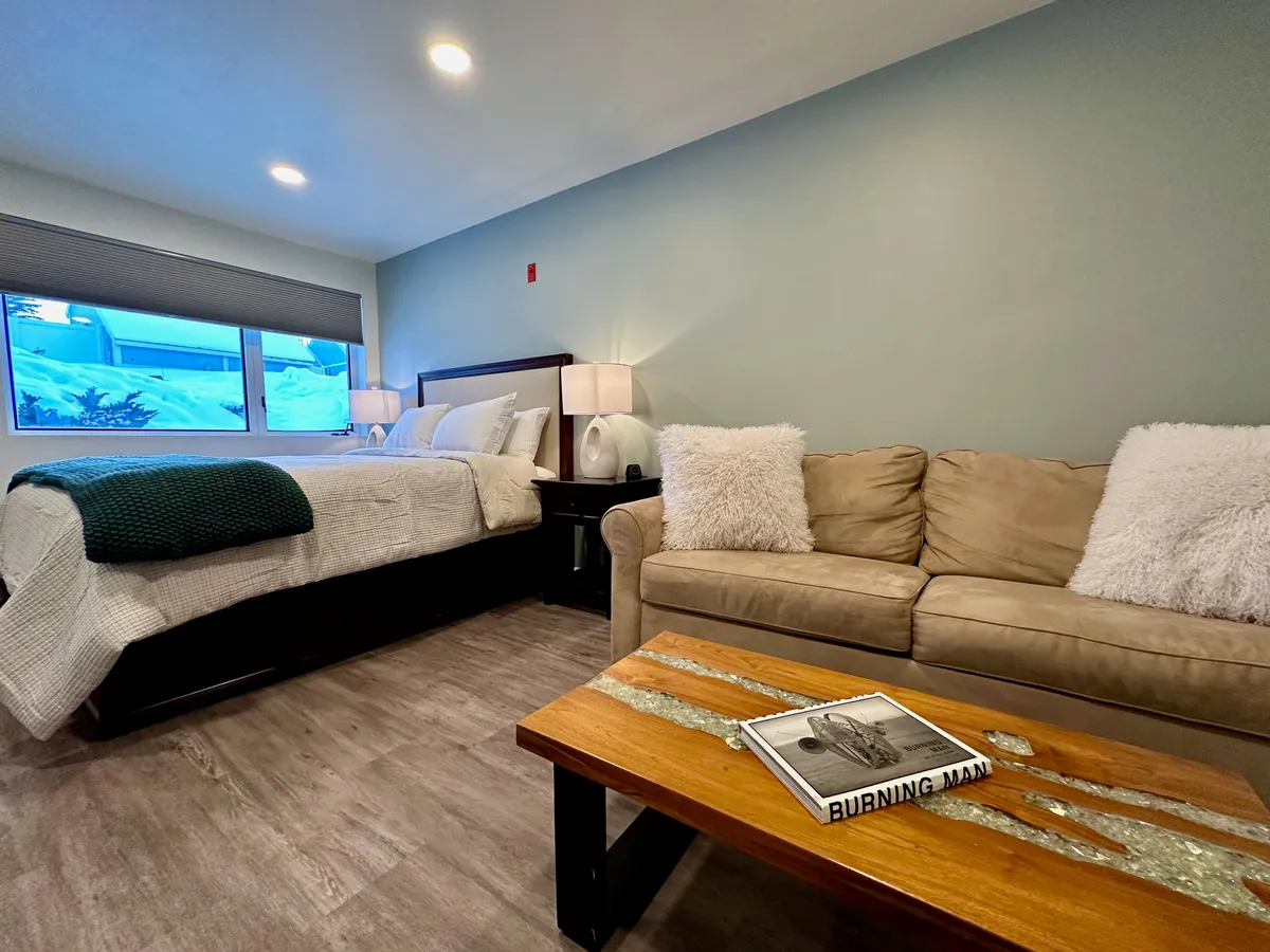 A cozy interior with a bed dressed in soft linens, a comfortable couch, and a wooden coffee table with a book atop it. The room is bathed in natural light and set up for both relaxation and casual snacking—ideal for enjoying breakfast in bed or gathering with friends.