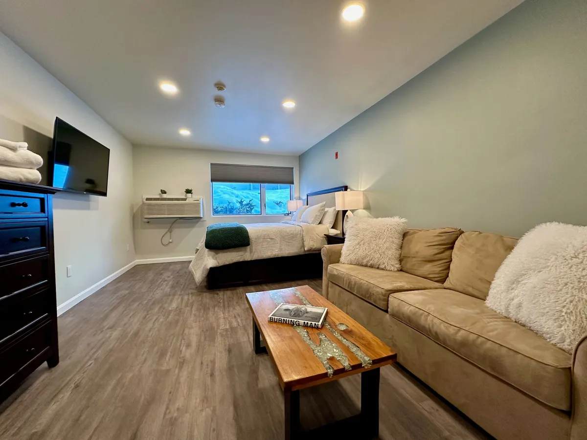 This inviting interior scene features both the plush queen bed and the comfy sofa, with a coffee table and Smart TV in view. The arrangement suggests a space perfect for relaxing, watching movies, or unwinding after a day of adventure. The window hints at natural light and a cozy atmosphere.