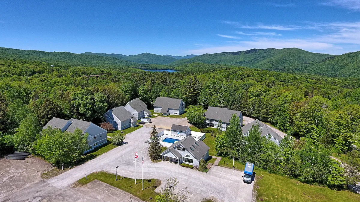 An expansive view of the main property building is seen alongside the swimming pool, set against a dramatic mountain backdrop. The parking area is visible in the foreground, with manicured lawns extending toward the pool and the mountains beyond. The serene environment suggests opportunities for family barbecues, outdoor events, and easy access to resort amenities.