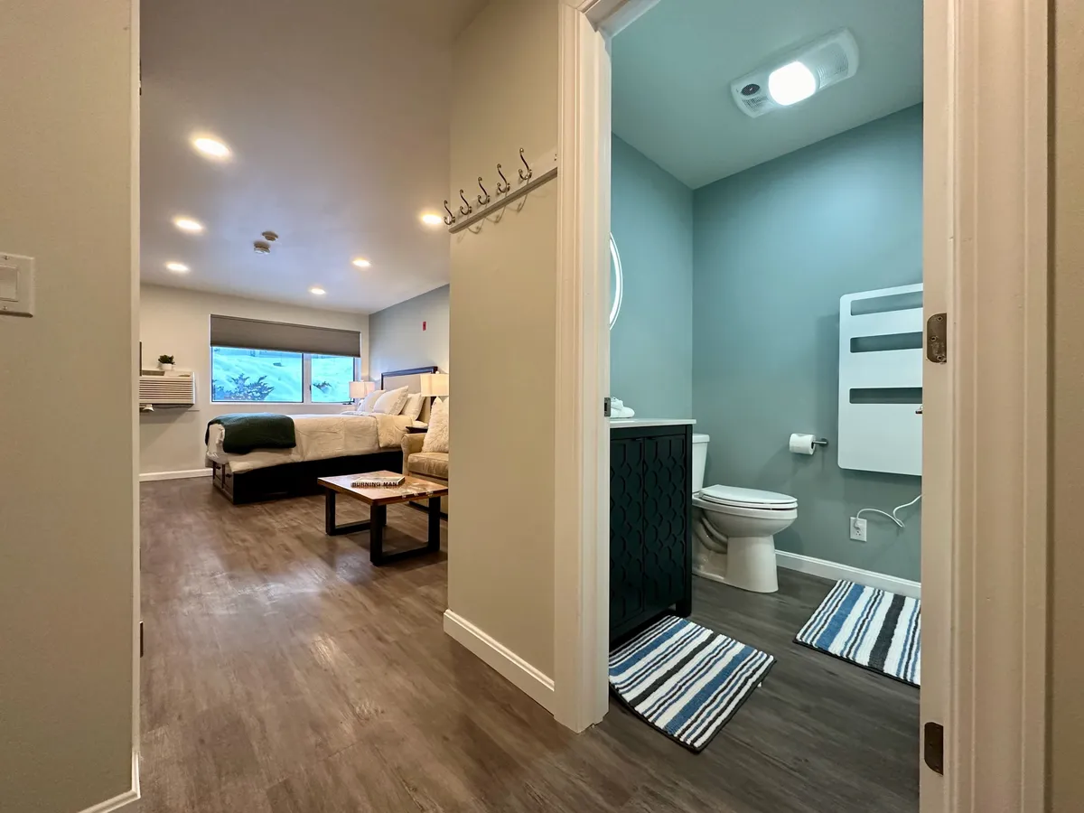 A cozy interior room with a bed draped in a green throw blanket, a small table with chairs positioned for conversation or a casual meal, and an entrance to the bathroom. Natural light filters in, adding a warm and welcoming ambiance. The space suggests comfort after a day outdoors and is ideal for quiet mornings or sharing stories over coffee.