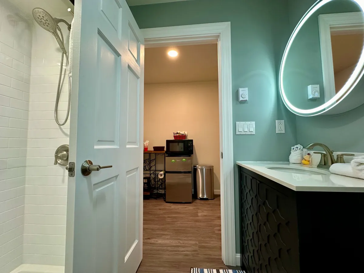 A sleek bathroom space with a glass shower enclosure, circular mirror above a vanity sink, and a view of the kitchenette access. The ambiance is clean, functional, and inviting.