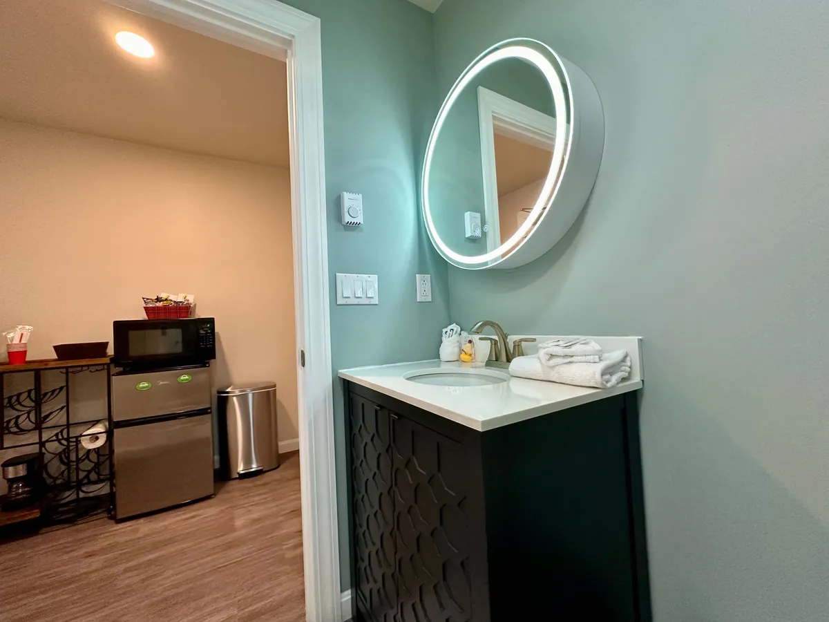 This scene captures a contemporary corner with a circular mirror, a modern sink, and neatly arranged towels, while a microwave and fridge are visible in the background. The lighting and décor create a functional yet stylish atmosphere, suggesting both comfort and convenience for short-term stays.