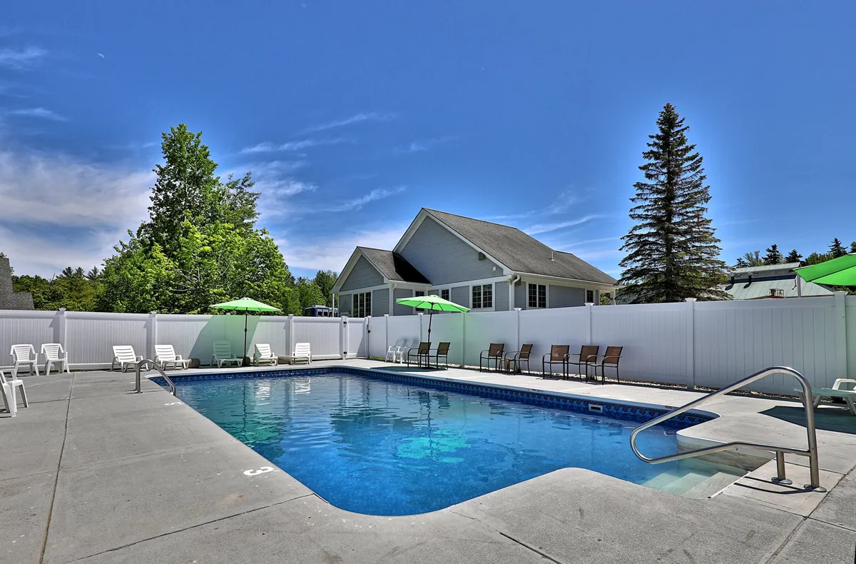 This image captures a vibrant outdoor swimming pool surrounded by green umbrellas and lounge chairs, with lush trees providing a tranquil backdrop. The area is inviting, with ample space for sunbathing, relaxing, or gathering with friends and family. The bright, cheerful atmosphere highlights the leisure-focused amenities available during the summer season.
