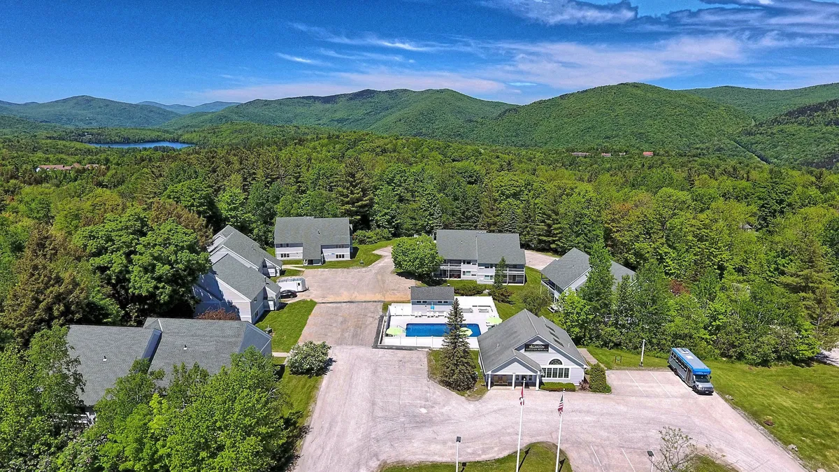 The image presents a peaceful exterior setting with a swimming pool, lush greenery, and the property buildings nestled beneath a stunning mountain backdrop. The scene captures the tranquility of poolside leisure and the allure of outdoor relaxation and family gatherings.